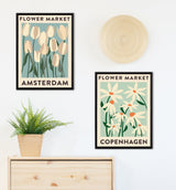 Two framed floral posters with black borders, one showing pale tulips labeled Amsterdam, the other white daisies labeled Copenhagen, hung above a potted fern and wooden furniture.