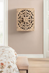 Light wood wall cabinet with symmetrical floral and vine carvings forming a circular pattern on two doors, mounted on a beige wall above a floral bedspread and bench.