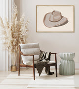 Minimalist framed canvas depicting a taupe cowgirl hat with shadow on a beige wall above a mid-century wooden armchair with light gray cushions, a textured blanket, and a green sculptural side table.