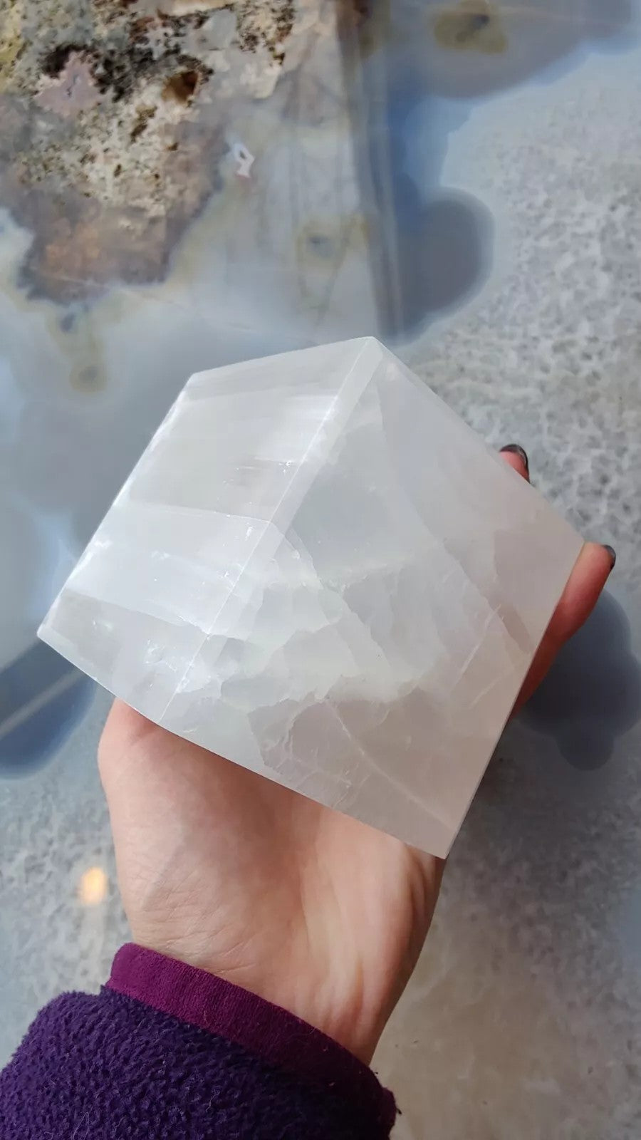 Hand holding a translucent white selenite cube with visible striations and smooth polished surfaces, set against a blurred glossy agate stone background.