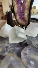 Three translucent white selenite cubes positioned on polished, multicolored agate slices with a purple and brown geode standing upright in the background.