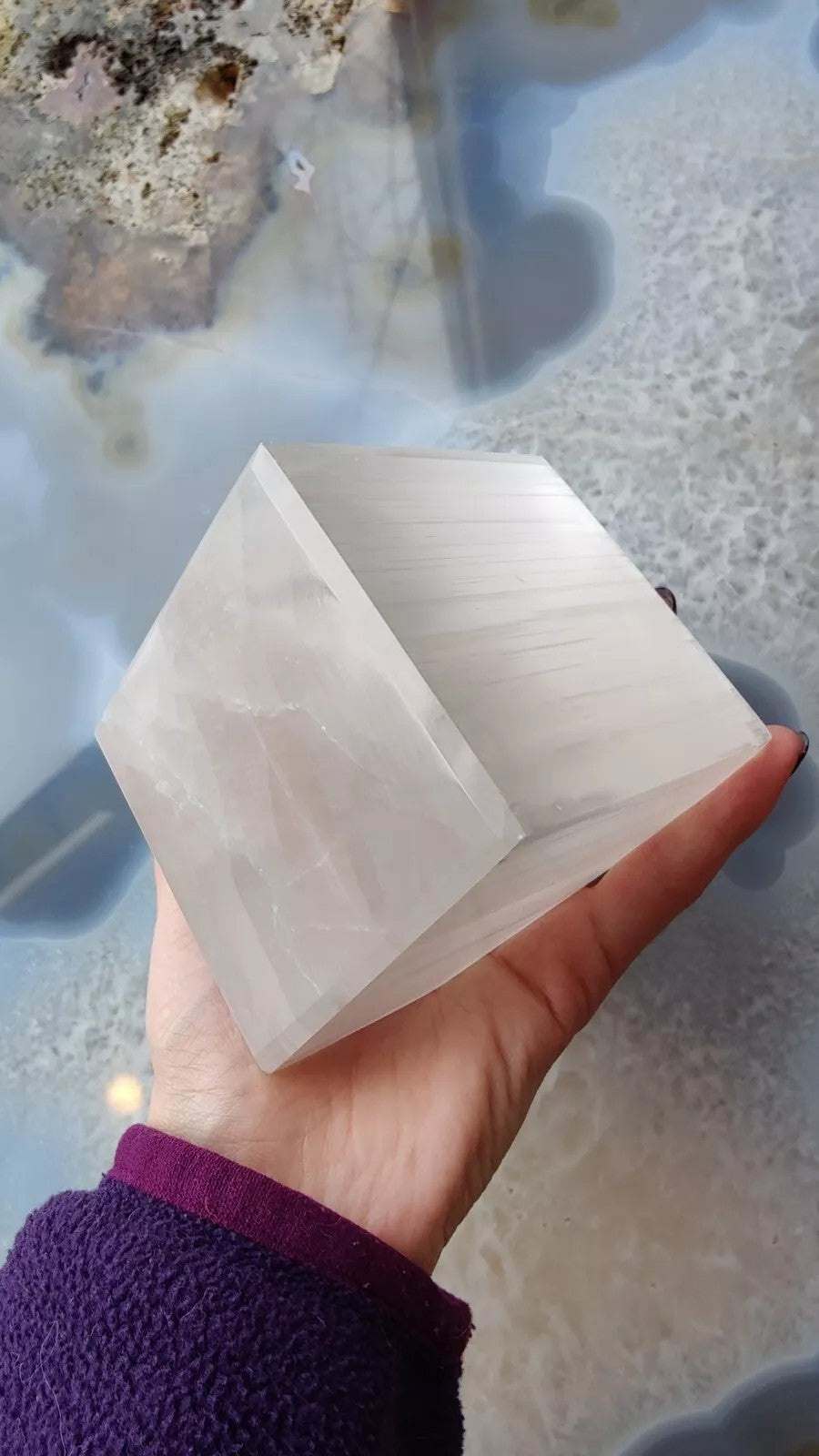 Hand holding a translucent selenite cube with smooth, polished faces and subtle horizontal striations, against a glossy blue and beige agate surface.