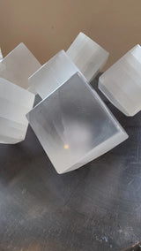Six translucent white selenite cubes with smooth, slightly frosted surfaces arranged irregularly on a gray textured surface against a beige background.