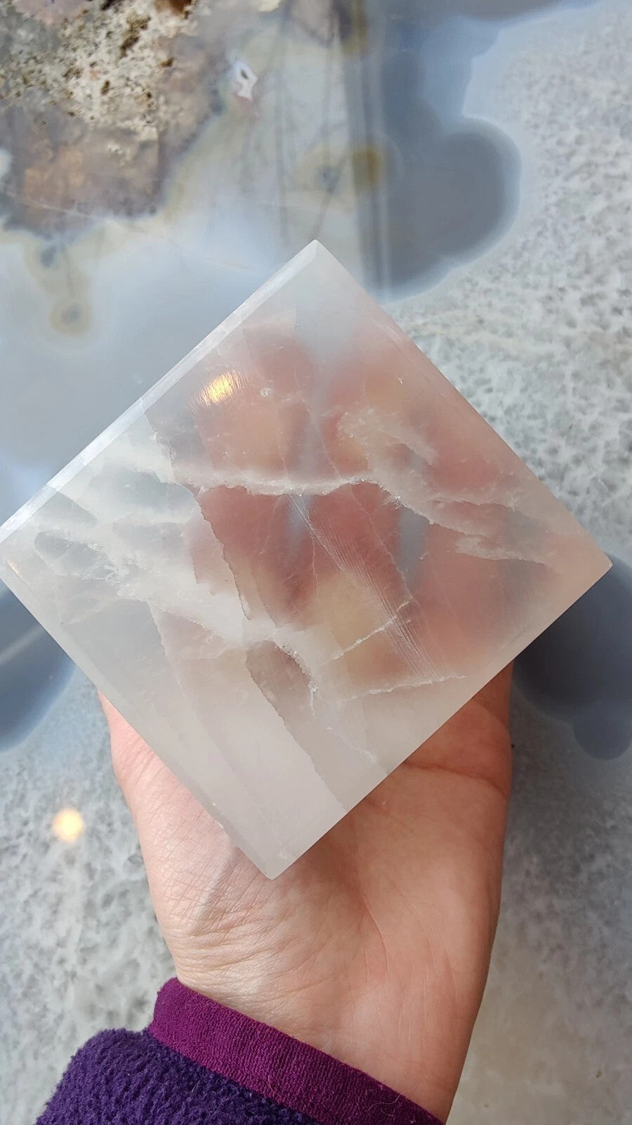 Translucent selenite cube with white and peach-colored internal striations held against a blue-gray patterned background.