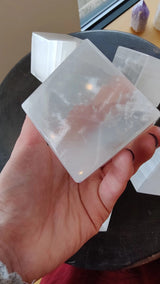 Hand holding a translucent white selenite cube with smooth, slightly cloudy surfaces reflecting light, a dark round tray and other crystals in the background.