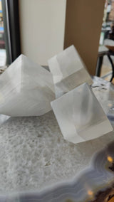Three translucent white selenite cubes with natural striations, positioned on a textured gray stone surface near a window and indoor setting.