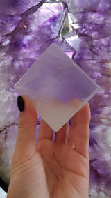 Hand holding a translucent, pale lavender selenite cube with beveled edges, set against a background of purple amethyst crystals with white and violet hues.