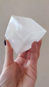 Hand holding a semi-translucent white selenite cube with smooth, beveled edges against a plain beige background.