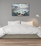 Framed canvas depicting a moonlit seascape with shining water, rocky shoreline, and distant hills, hanging above a white bed against a gray wall.
