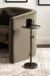 Polished dark metal adjustable table with a round top and base, holding a black mug, placed on a patterned beige carpet beside a curved olive green sofa.