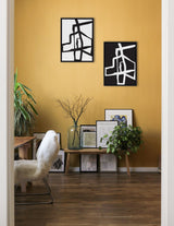 Two framed abstract black and white canvases with bold intersecting strokes hang on a warm yellow wall above a wooden bench holding various art prints and plants.