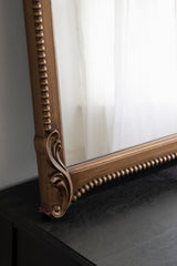 Close-up of a bronze-toned arched wall mirror with decorative beading and carved scroll detail on its wooden frame, resting on a dark surface.