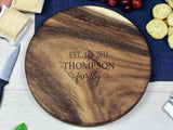 Small round wood charcuterie board with rich grain, engraved with 'EST. 2011 THOMPSON family' centered, shown with cheese, crackers, grapes, and a knife on a light wood table.