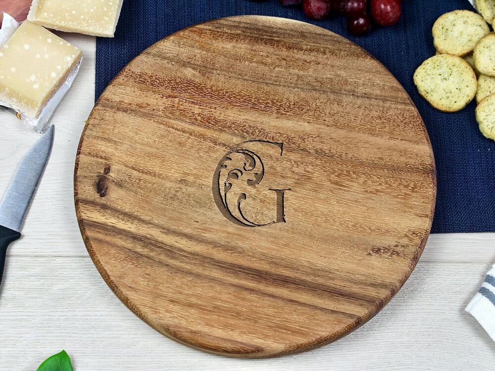 Small round wooden charcuterie board with a carved letter G and decorative flourish at the center, resting on a light wood surface with cheese, grapes, crackers, and a knife nearby.