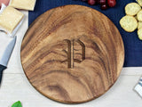 Round wooden charcuterie board with natural grain pattern and engraved letter P in the center, placed on a light surface with cheese, grapes, and crackers nearby.