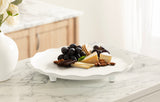 White scalloped round tray with three short legs holding grapes, assorted cheeses, almonds, and crackers on a marble countertop near a wooden cabinet and window.