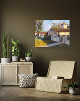 Canvas print of a sunlit suburban street scene with houses, trees, and a white fence, displayed on a brown wall above a wicker chair and potted plants.