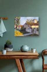Impressionist painting of houses and trees with sunlit yellow and brown tones, displayed on a muted green wall above a wooden table with a gray teapot, two cups, and a black and white plant pot.
