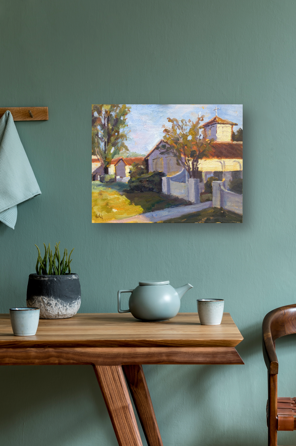 Impressionist painting of houses and trees with sunlit yellow and brown tones, displayed on a muted green wall above a wooden table with a gray teapot, two cups, and a black and white plant pot.