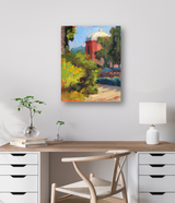 Colorful impressionist painting of a red tower with a white dome, surrounded by green foliage and a sunlit path, displayed above a wooden desk with a lamp and plant.