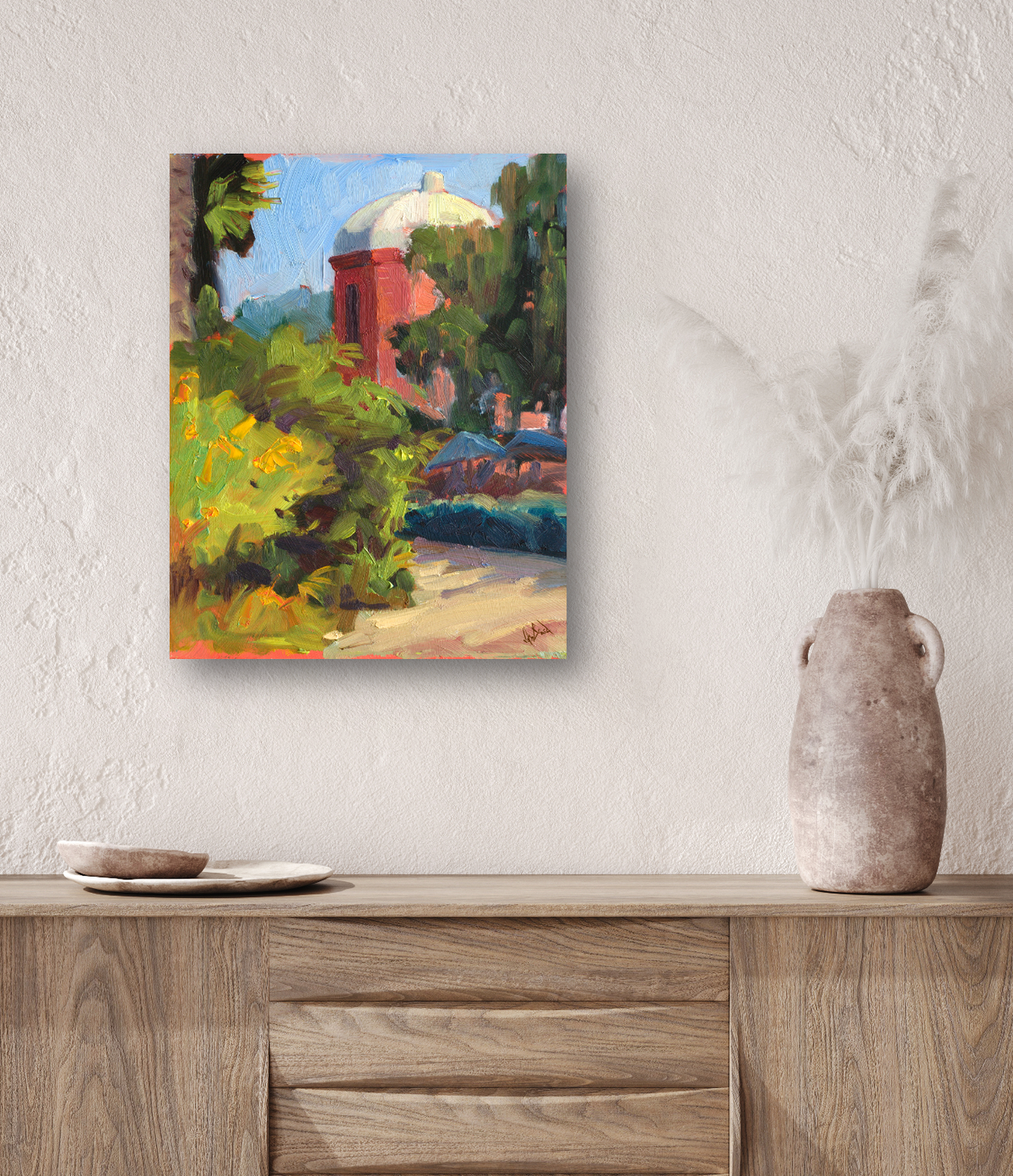 Colorful impressionist painting of a red tower with a white dome partially obscured by green and yellow foliage, mounted on a textured white wall above a wooden sideboard.