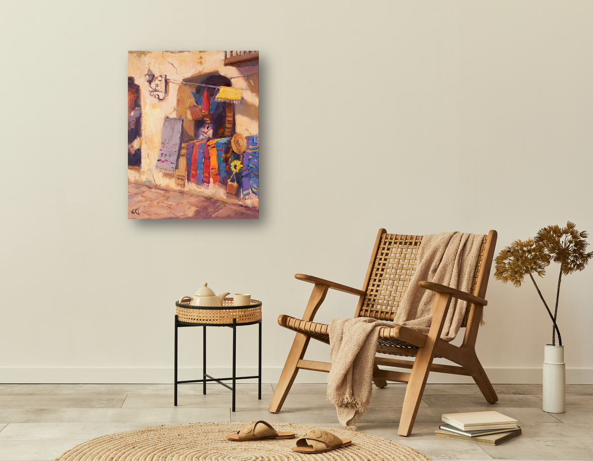 Colorful Sarapes Enhanced Canvas Print depicting vibrant fabric market scene, hanging on a beige wall above woven chair and small black table with pottery in cozy corner