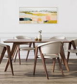 Rectangular wooden dining table with angled legs surrounded by four cream upholstered chairs, beneath a framed abstract landscape painting in soft pastel tones.