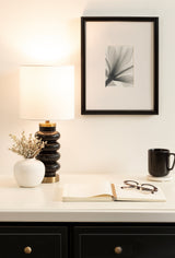 Black Rogala table lamp with stacked rounded base and white drum shade on white surface next to small white vase with delicate flowers, open book, glasses, and black mug under framed abstract art.