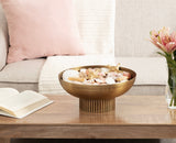 Round bronze ribbed metal bowl filled with seashells, placed on a wooden coffee table with a beige sofa and pink cushion in the background.