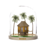 Miniature tropical hut with patterned walls and a thatched roof, surrounded by three palm trees and a rope hammock under a clear dome on a round base.
