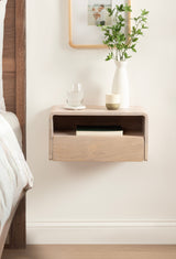 Light wood floating side table with a drawer and open shelf holding a book, topped with a white vase of greenery and a glass of water, beside a bed with white linens.