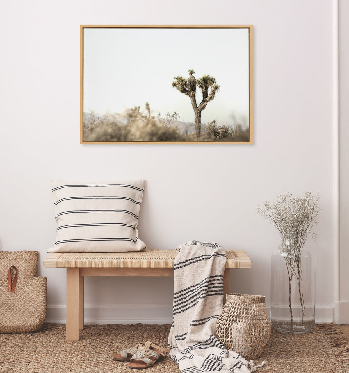 Minimalist interior with a wooden bench, striped pillow and blanket, woven baskets, and a framed canvas of a single Joshua tree against a pale sky on the wall.