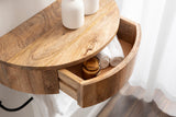 Round floating side table made of warm-toned wood with a visible drawer containing rolled white towels and a brown glass jar, against sheer white curtains.