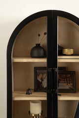 Tall, black-framed arched wall cabinet with glass doors containing a black vase with dried stems, framed photos, and a white candle in a brass holder.