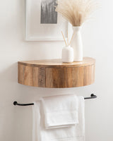 Curved floating wood side table with a warm brown grain mounted on a white wall, topped with white vases holding dried grasses and reed diffusers above a black towel bar with folded white towels.