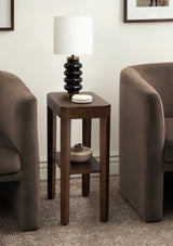 Tall dark wood side table with a lower shelf holding a beige ceramic bowl, topped by a black segmented lamp with a white drum shade between two taupe upholstered chairs.
