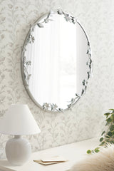 Oval wall mirror with metal frame adorned by delicate floral and leaf accents in muted silver, mounted on light floral patterned wallpaper.