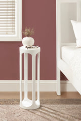 Tall, slender white side table with elongated oval cutouts stands on a beige rug beside a white bed frame against a muted red wall.