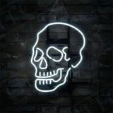 White neon light shaped like a human skull with prominent eye sockets and teeth, mounted on a dark brick wall with mounting hardware visible.