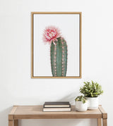 Framed canvas art of a tall green cactus with a large soft pink flower on top, hanging above a wooden table with two potted plants and stacked books