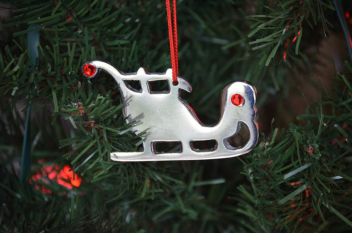 Silver metal sleigh Christmas ornament with two red gem accents, hanging by a red string on a green pine tree branch.