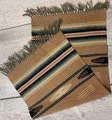Small woven kilim rug with earthy brown base, featuring horizontal stripes in cream, black, red, and green, and fringed edges on a light wood floor.