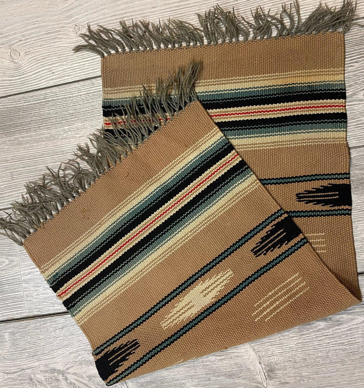 Small woven kilim rug with earthy brown base, featuring horizontal stripes in cream, black, red, and green, and fringed edges on a light wood floor.
