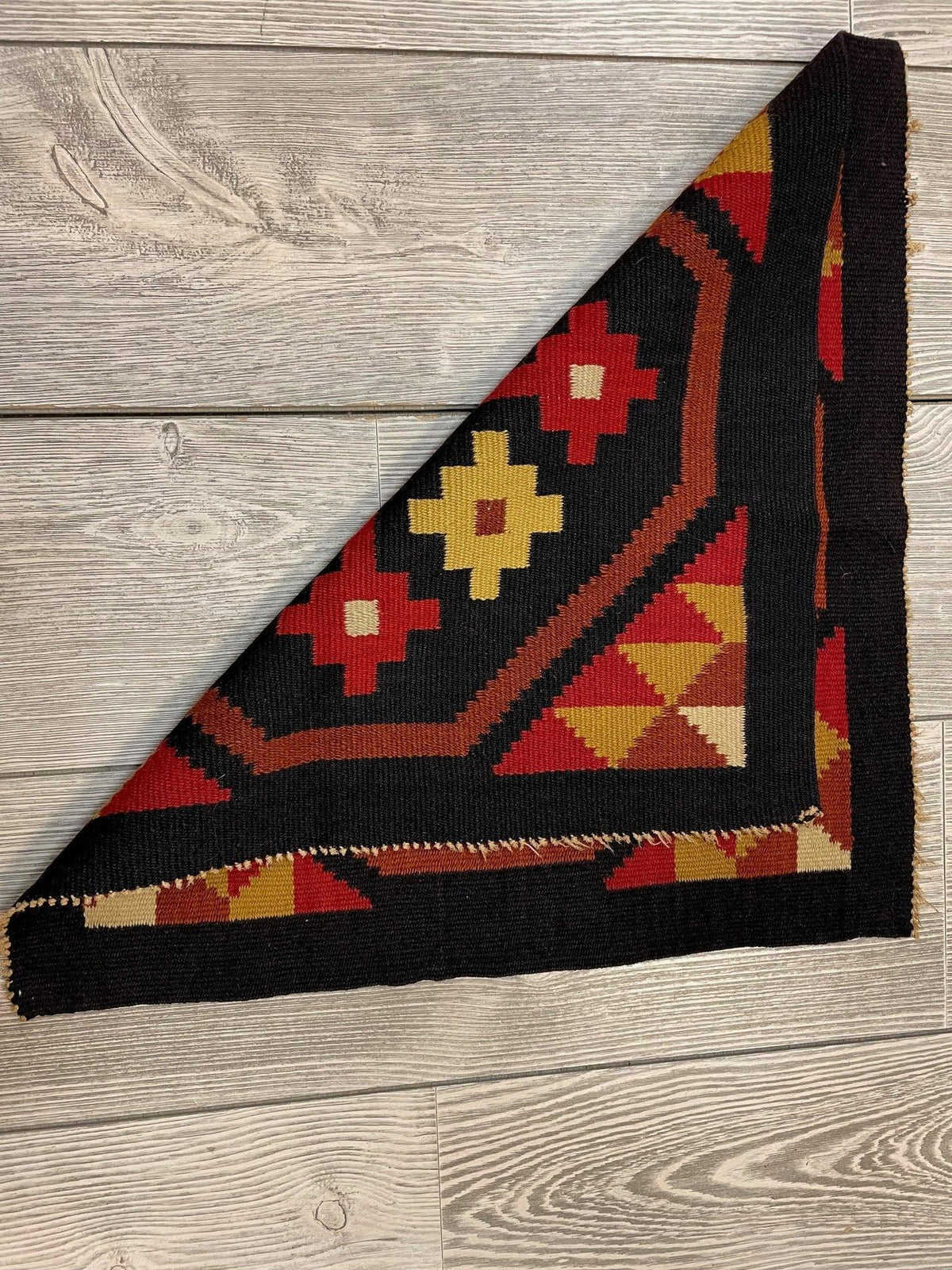 Small square Mexican hand woven kilim rug in red, yellow, black, and tan geometric floral patterns, folded to show both front and back sides on wood floor.