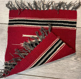Small red hand-woven Mexican Kilim rug with black and cream stripes near the fringed edges, shown partially flipped on a light wood floor.