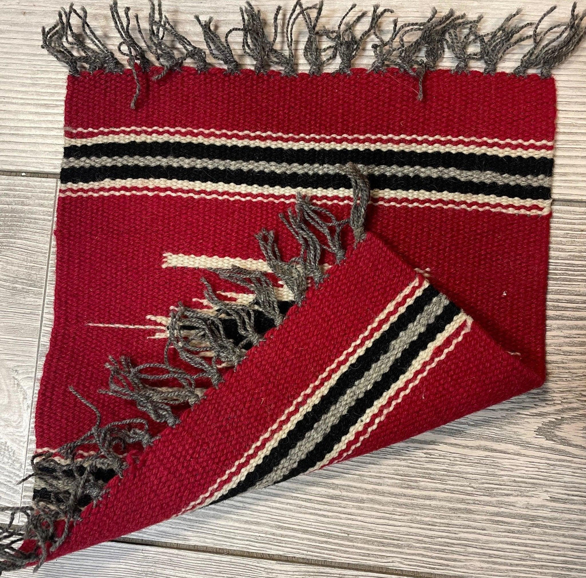 Small red hand-woven Mexican Kilim rug with black and cream stripes near the fringed edges, shown partially flipped on a light wood floor.