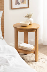 Round wooden side table with three legs and lower shelf holding books, topped with a white vase of dried flowers and a small bowl, next to a bed and window.