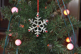 Silver snowflake ornament with red gem accents hanging on a green Christmas tree with round pink string lights in the background.