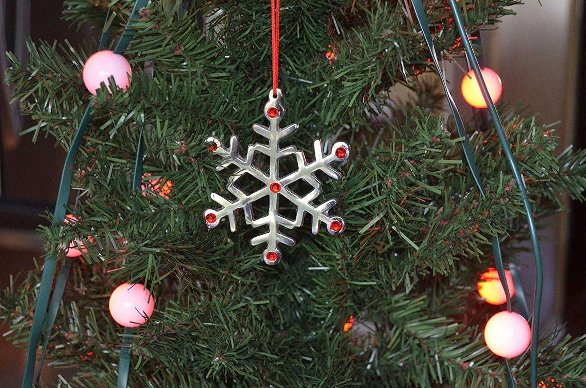 Silver snowflake ornament with red gem accents hanging on a green Christmas tree with round pink string lights in the background.