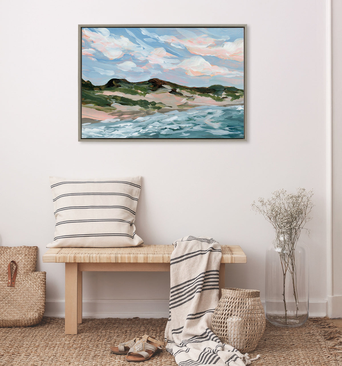Framed canvas painting of a beach scene showing blue water, sandy dunes with green vegetation, and a sky with pink and blue clouds, hanging above a light wood bench with a striped pillow and throw.